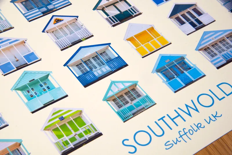 Close-up of the Southwold print at an angle: rows of cut-out beach huts on cream paper, with the word Southwold and the handwritten line Suffolk UK