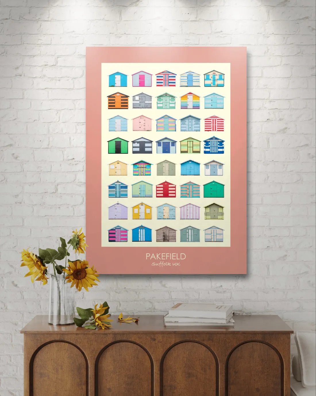 The Pakefield print with a coral border on a white brick wall above a walnut sideboard, with sunflowers in a vase and a stack of books