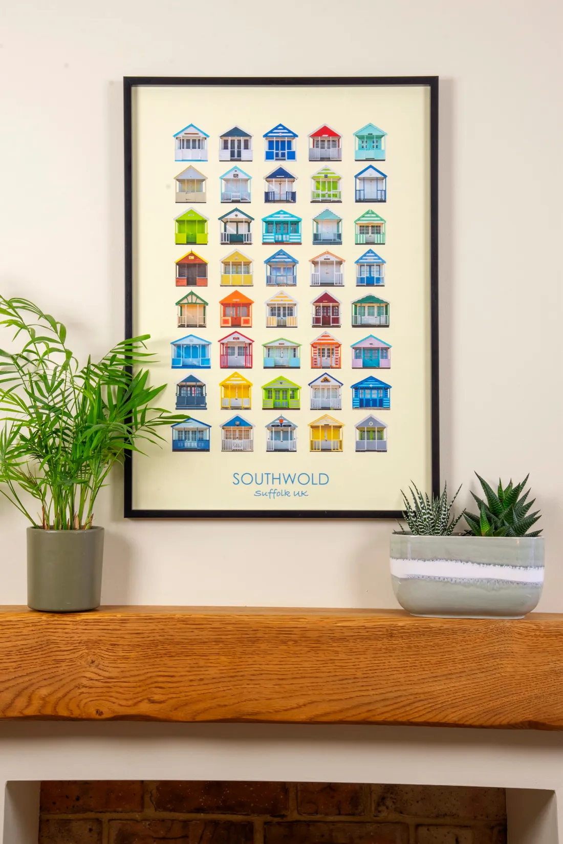 The Southwold print in a slim black frame on a cream wall above an oak mantel shelf, between a palm and a succulent in a striped pot