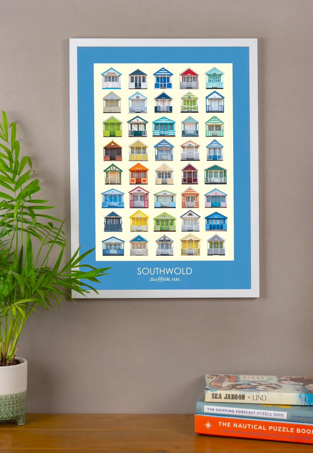 The Southwold print with a True Blue border in a white frame on a grey wall, above a stack of nautical books and a plant